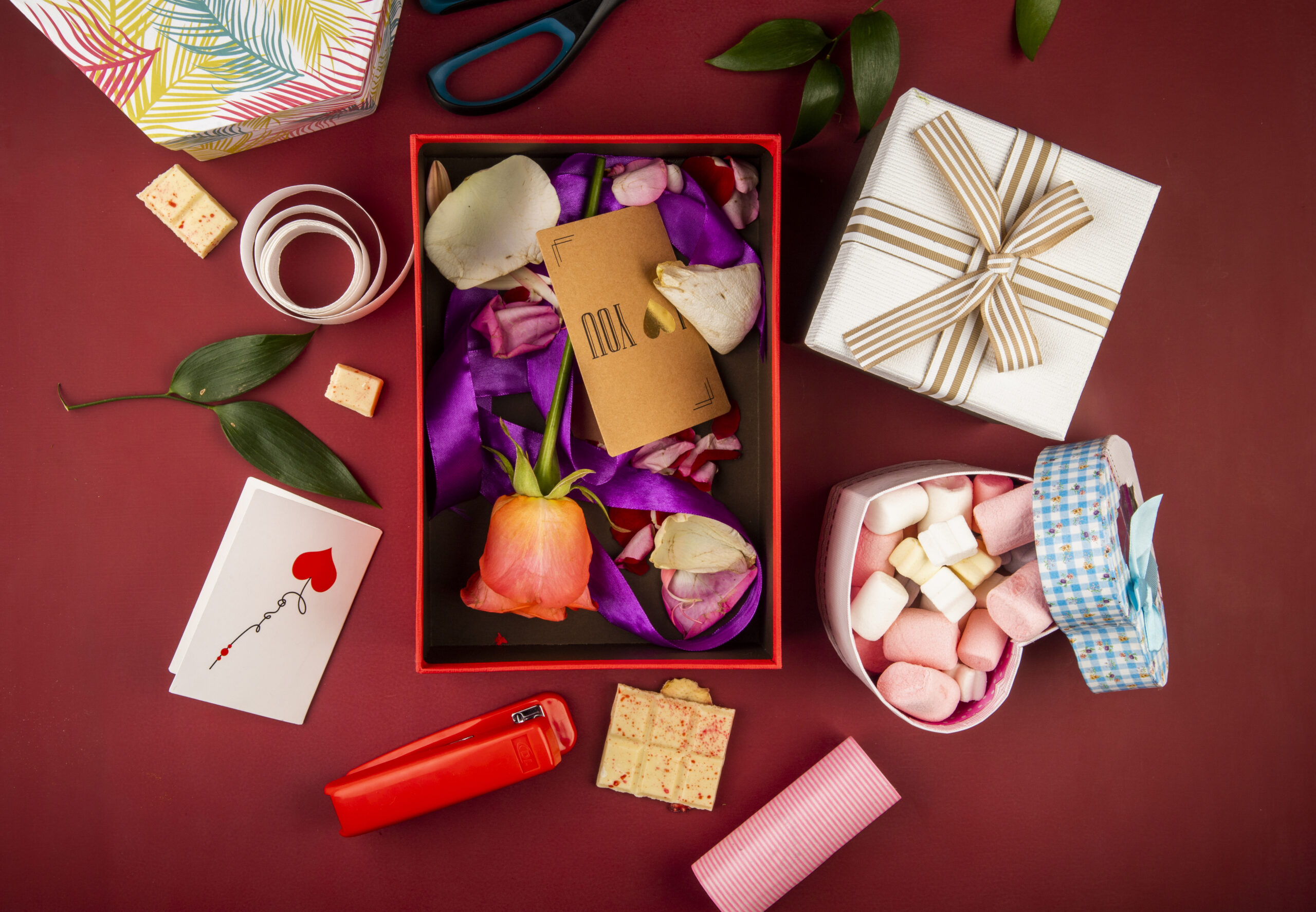 top view of a red present box with brown paper card and coral color rose flower and petals with purple ribbon and heart shaped box filled with marshmallow on dark red background