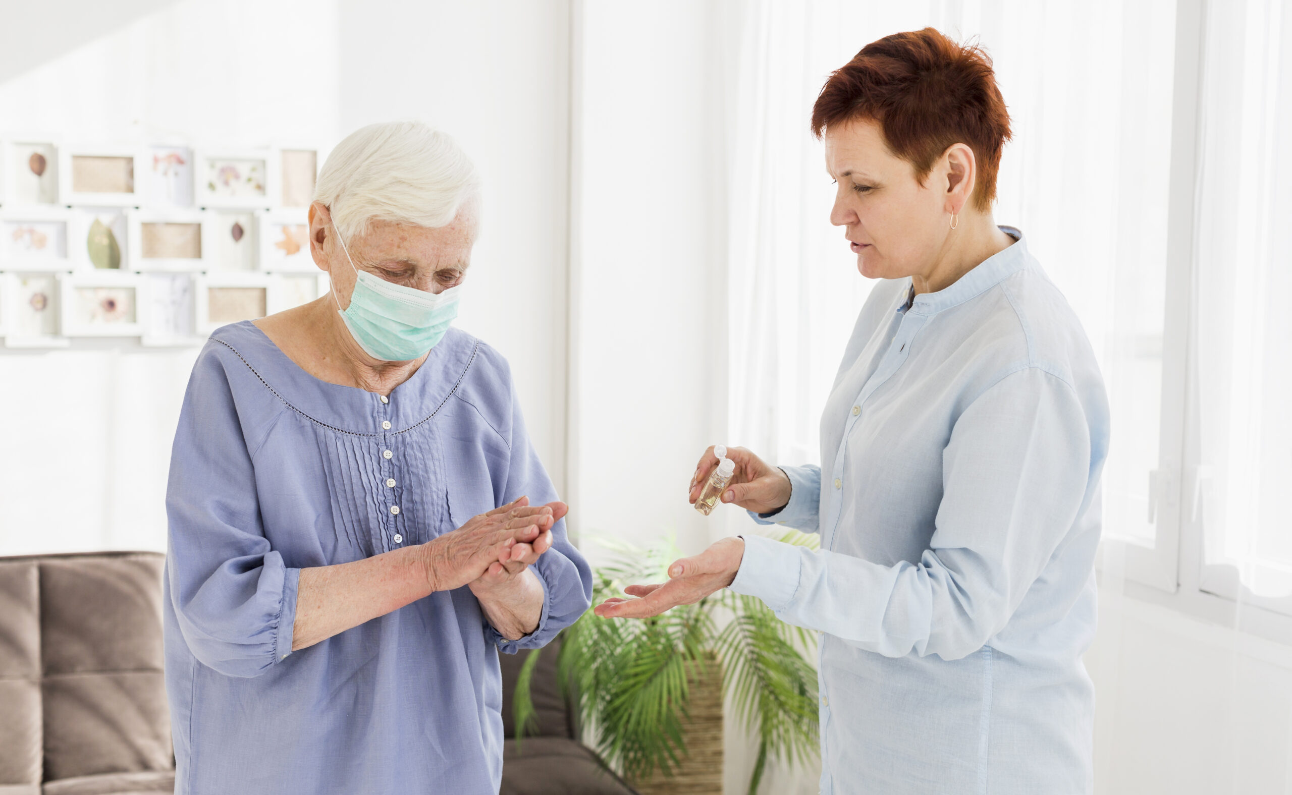 Older woman wearing a face mask sanitizes her hands while another woman dispenses hand sanitizer in a bright living room.